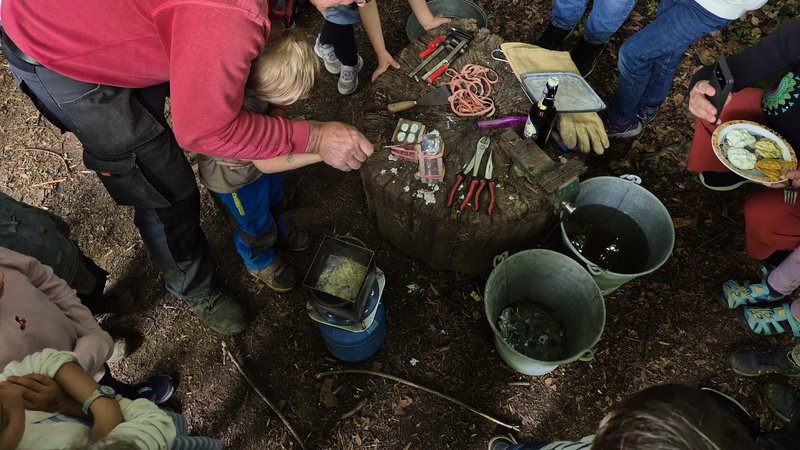 Zinngießen am Sommerfest: Gussformen und Werkzeuge auf einem Baumstumpf, Kinder schauen zu.